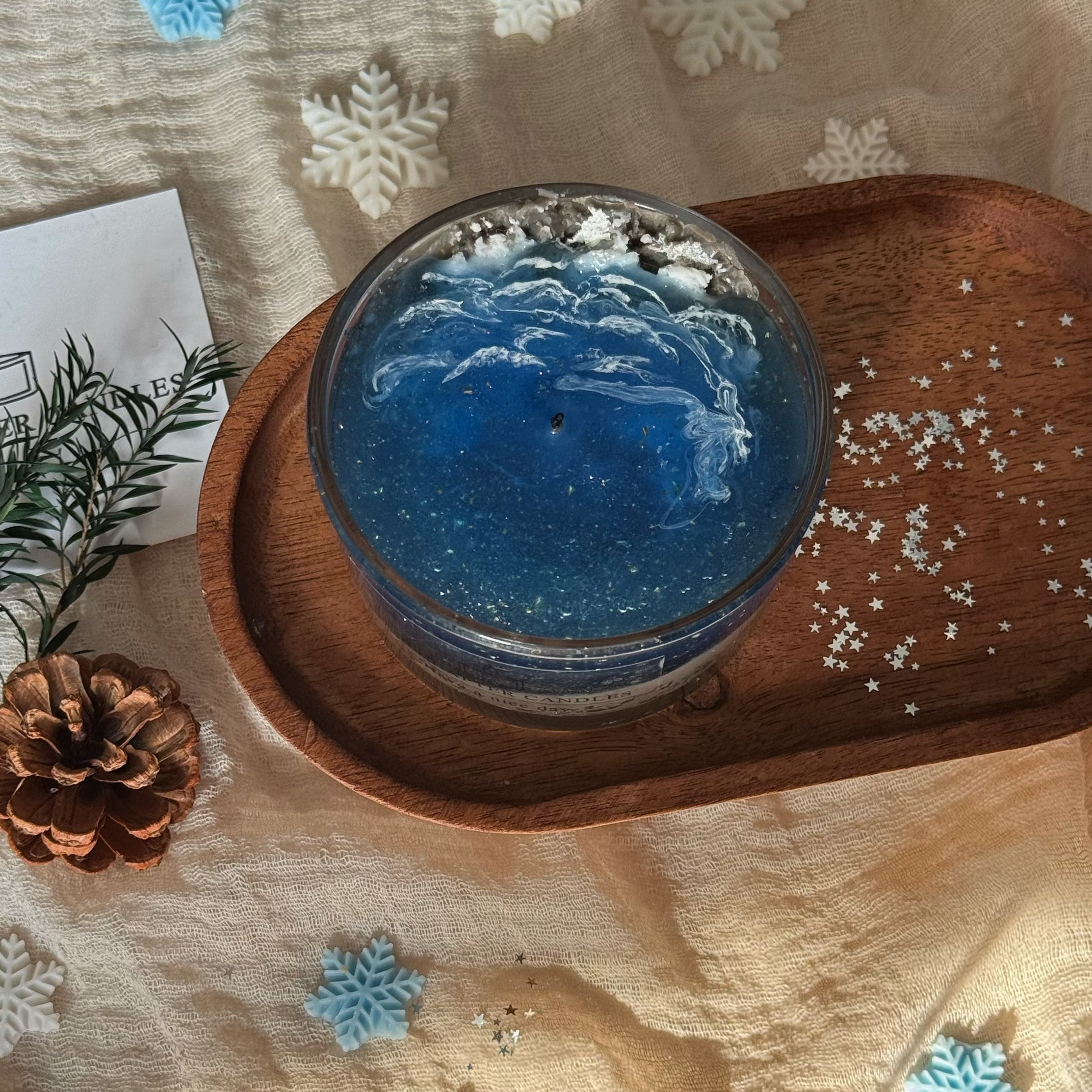Relaxing home decor setting showcasing an exquisite transparent blue sea sachet candle with a branded label "OUR CHAPTER CANDLES". Warm natural candlelight on soft fabric for bedroom aromatherapy.