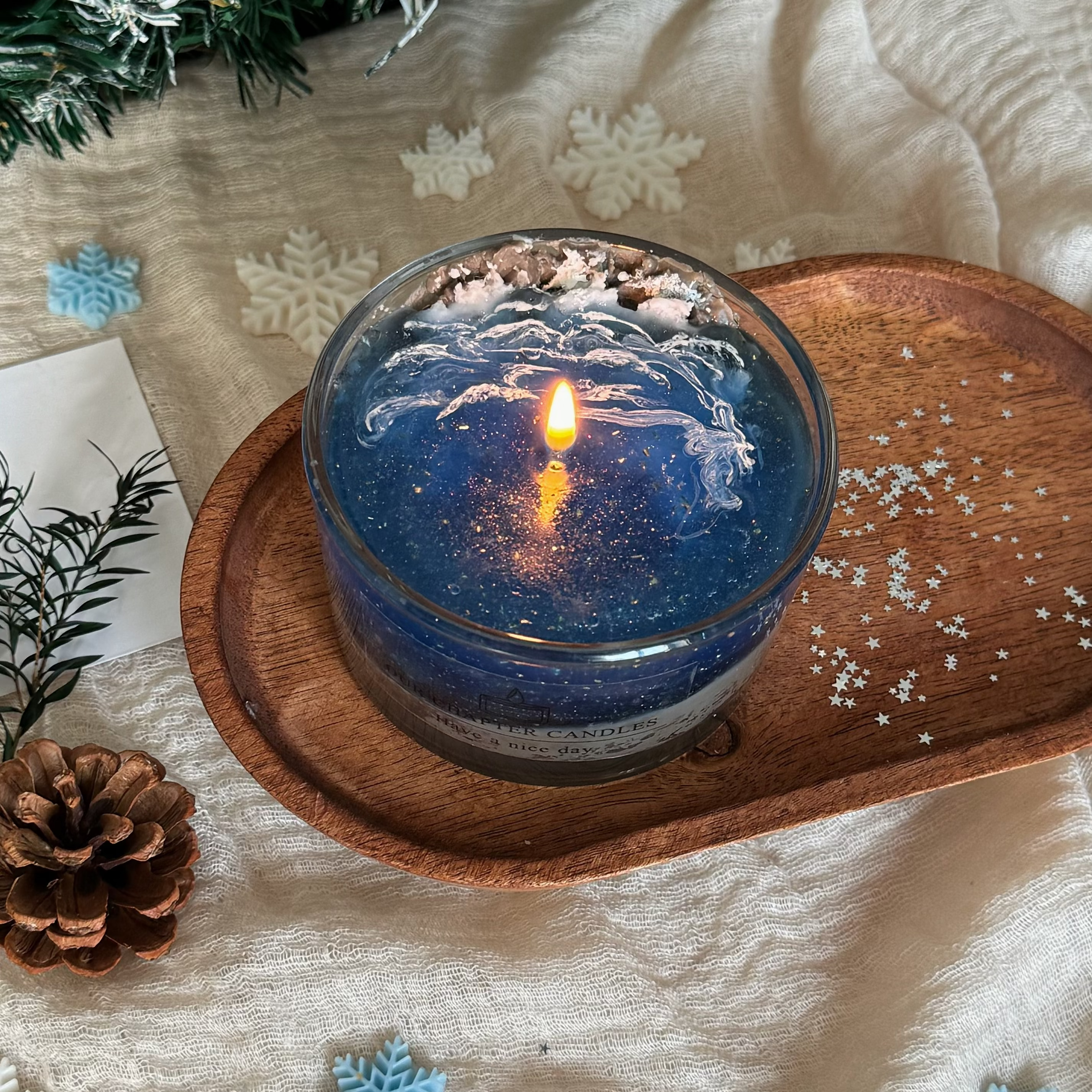 A lit luxury ocean wave jelly candle glowing with a warm flame based on image_0fbffb.jpg. The deep blue gel wax features starry glitter and detailed white foam waves, resting elegantly on a rustic wooden tray.