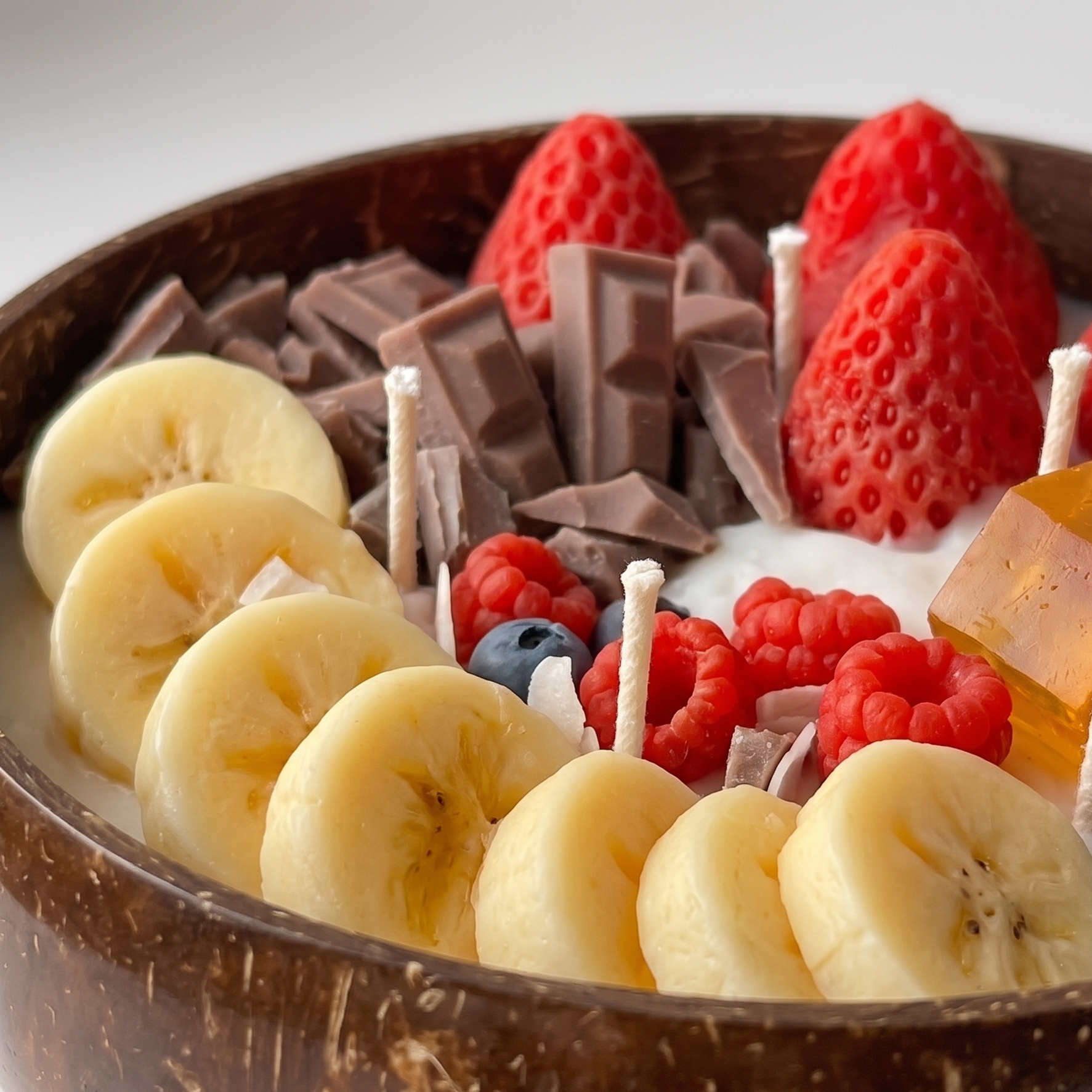 Extreme macro close-up of a luxury dessert candle showing detailed textures of realistic wax strawberries, banana slices, and the fibrous organic surface of a natural coconut bowl.