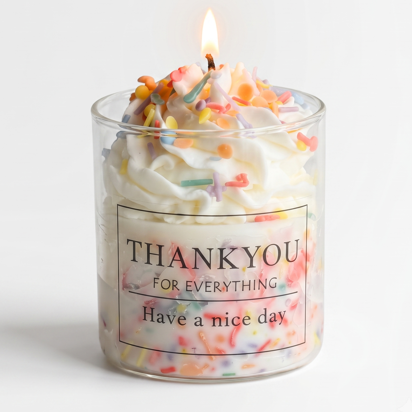 A burning luxury cupcake dessert soy wax candle in a clear glass jar topped with whipped white cream wax and colorful candy sprinkles. Features a minimalist thank you gift label on a pure white background.