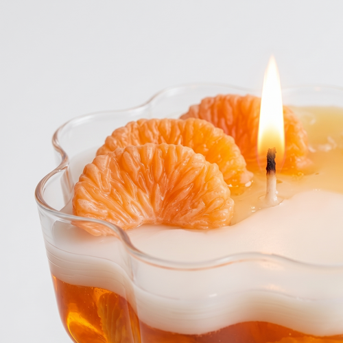 Extreme macro close-up of a lit dessert candle showcasing the detailed waxy texture of realistic mandarin orange segments and a glowing flame. High-resolution view of the wavy white soy wax layer inside a petal-shaped coupe glass.