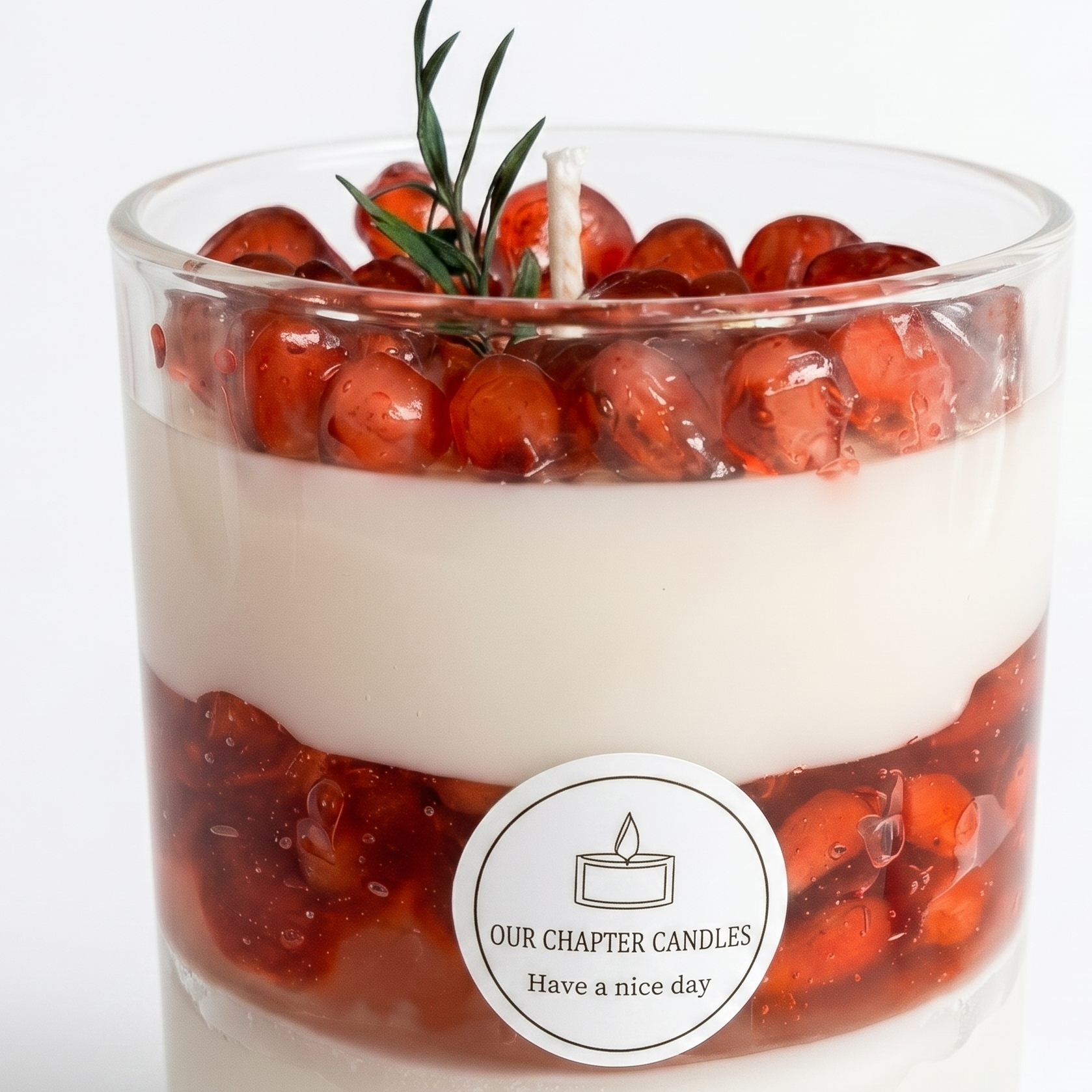 Extreme macro close-up of a dessert candle showcasing the glossy waxy texture of ruby pomegranate seeds and a green herbal sprig. High-resolution view of the multi-layered soy and gel wax base with a sharp circular brand label.